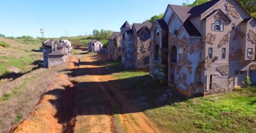 Abandoned houses line a dirt road, surrounded by overgrown land and clear blue skies.