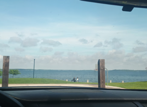 View of a lake from inside a car, with two people sitting on a bench by the water under a partly cloudy sky.