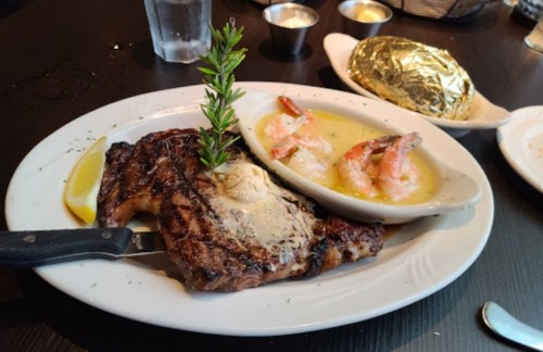 A grilled steak with a sprig of rosemary, served with shrimp and a baked potato on a white plate.