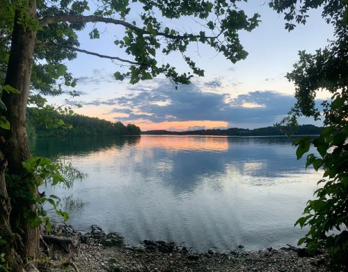 Serene lake view at sunset, framed by trees, with calm water reflecting the colorful sky and distant shoreline.
