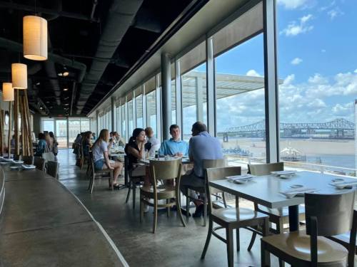 A modern restaurant interior with diners enjoying meals, large windows showcasing a river and bridge view.