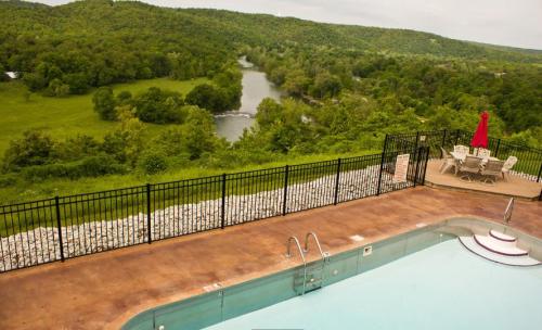 A scenic view of a pool overlooking a lush green landscape and a winding river, with a patio and red umbrella nearby.