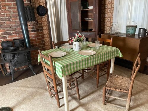 A rustic dining area with a green checkered tablecloth, wooden chairs, and a vintage stove in a cozy setting.