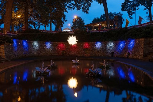 A serene pond at dusk, illuminated by colorful lights, with swans and a star-shaped decoration reflecting in the water.
