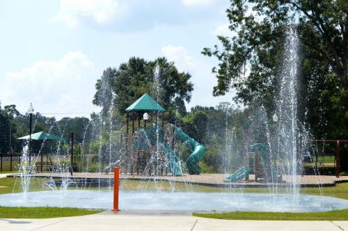 A splash pad with water jets in front of a playground featuring slides and climbing structures under a blue sky.