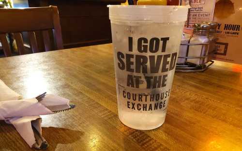 A clear cup with the text "I GOT SERVED AT THE COURTHOUSE" on a wooden table, with utensils nearby.