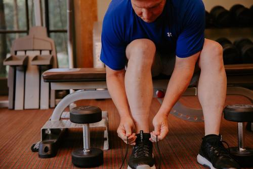 A person in a blue shirt ties their shoelaces while sitting on a bench in a gym. Dumbbells are visible in the foreground.