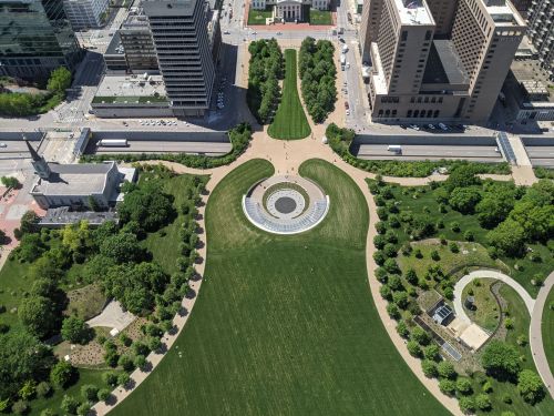 Aerial view of a green park with circular pathways, surrounded by buildings in an urban setting.