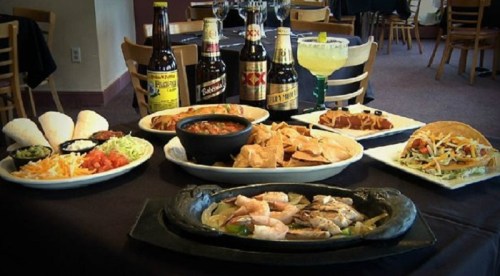 A table set with various Mexican dishes, including shrimp, tacos, nachos, and drinks, in a restaurant setting.
