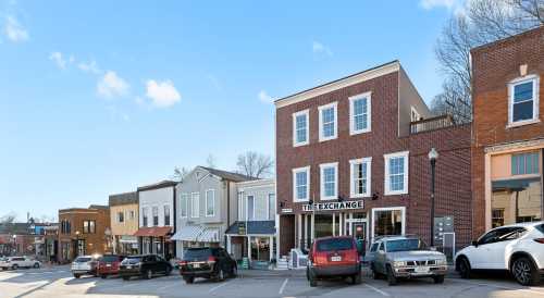 A quaint street scene featuring a mix of brick and colorful storefronts, with parked cars and a clear blue sky.