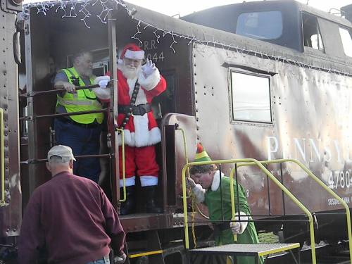 Santa Claus waves from a train car, while an elf and two men stand nearby, decorated with holiday lights.