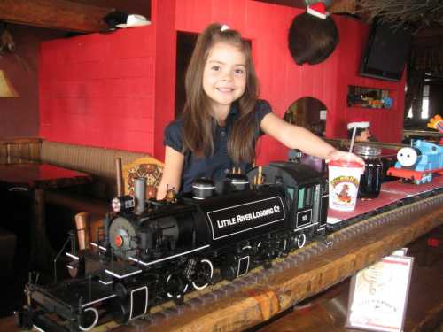 A smiling girl stands beside a model train on a table, holding a drink in a cozy, rustic setting.