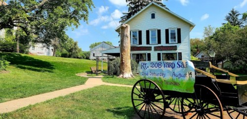 A historic house with a yard sale sign, a horse-drawn carriage in the foreground, and green grass under a blue sky.