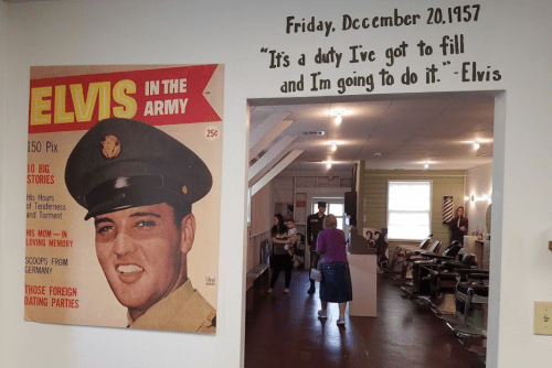 A vintage Elvis magazine cover hangs on a wall, with a quote above it and people in a room in the background.