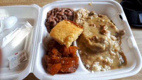 A takeout container with rice, brown gravy, beans, cornbread, and a side of sweet potatoes.