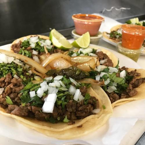 Three tacos filled with seasoned meat, onions, and cilantro, served with lime wedges and two sauces on the side.