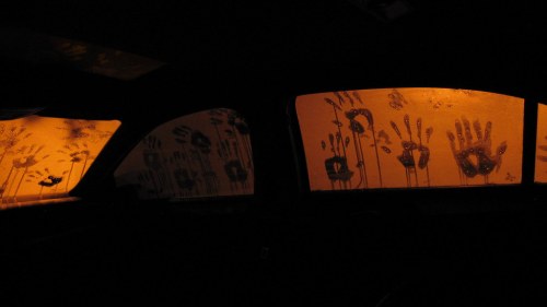 Foggy car windows with handprints and an orange glow from outside, creating a haunting atmosphere.