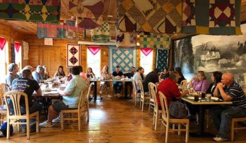 A cozy restaurant interior with wooden walls, quilted decorations, and groups of people dining at tables.