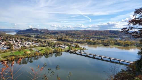 A scenic view of a river surrounded by hills and trees, with a bridge connecting two land areas under a blue sky.