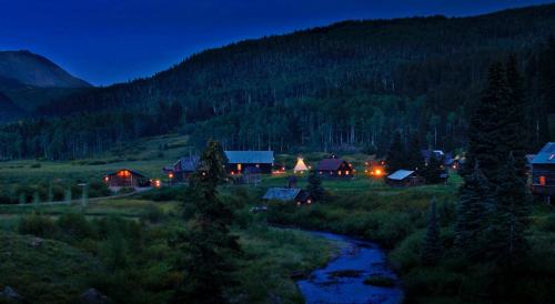 A serene village at dusk, with glowing lights and a winding stream, surrounded by mountains and lush trees.