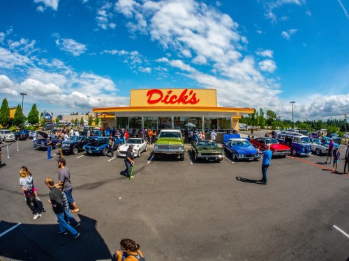 A vibrant car show outside a Dick's store, featuring classic cars and a crowd enjoying the event under a blue sky.