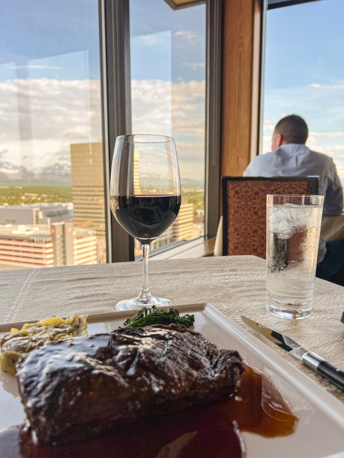 A gourmet meal with steak, wine, and a city view, while a person sits in the background, gazing out the window.