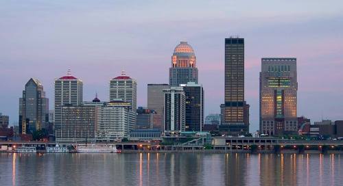 Skyline of a city at dusk, featuring tall buildings reflected in a river with a soft pink and blue sky.