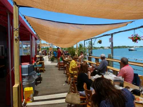 Outdoor dining area by the water, with people enjoying meals under shade sails and a scenic view of the bay.