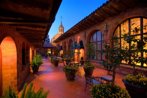 A serene courtyard with potted plants, stone arches, and a church tower in the background, illuminated at dusk.