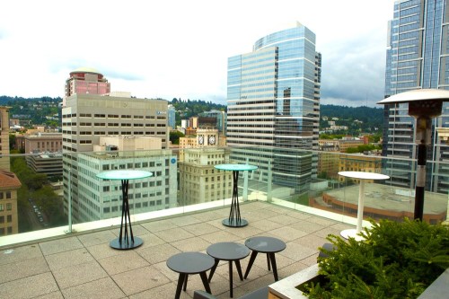 Rooftop view of a city skyline with modern buildings, glass tables, and seating on a cloudy day.