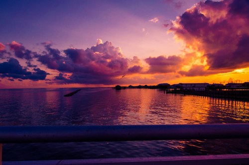 A vibrant sunset over calm waters, with colorful clouds reflecting on the surface and a pier in the distance.