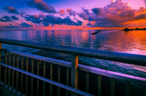 A vibrant sunset over calm waters, with colorful clouds reflecting on the surface, viewed from a pier railing.