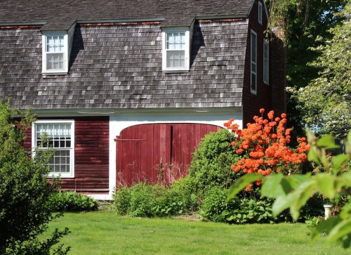 A charming red barn with a shingled roof, surrounded by vibrant orange flowers and lush greenery.