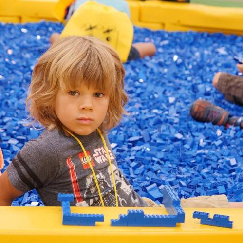 A child with curly hair sits in a pool of blue LEGO bricks, looking curiously at the camera.
