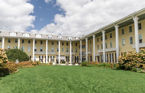 A sunny courtyard of a yellow building with columns, surrounded by green grass and blooming flowers under a cloudy sky.