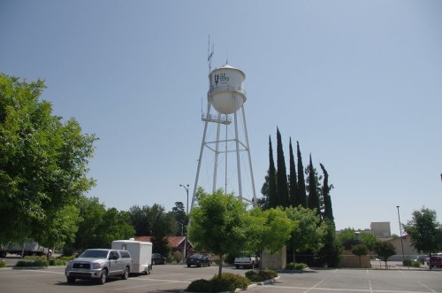 A tall water tower stands in a parking lot, surrounded by trees and clear blue skies.