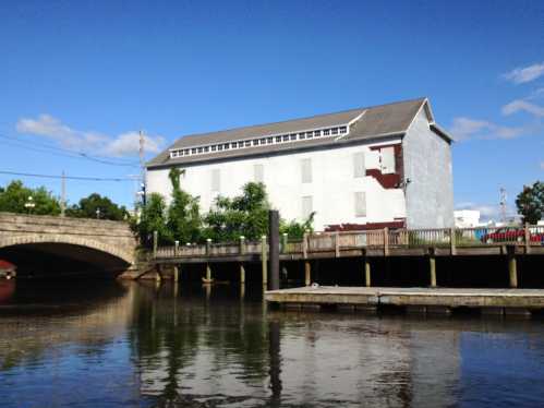 A large, white building by a river, with a bridge in the foreground and blue sky above.