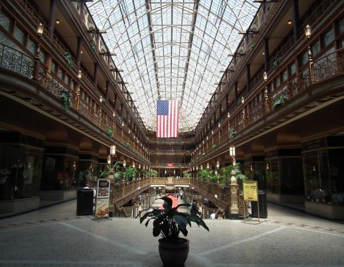Interior view of a grand atrium with a glass ceiling, featuring an American flag and lush greenery.