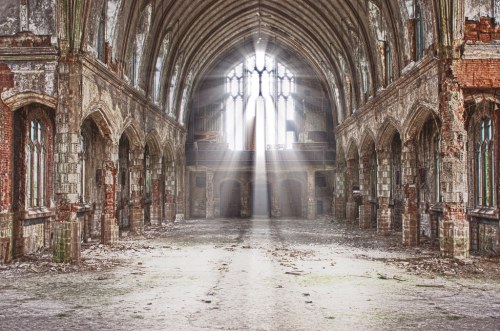 Abandoned church interior with crumbling walls, tall arches, and sunlight streaming through stained glass windows.