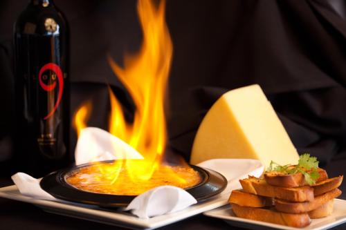 A flaming dish on a black tablecloth, accompanied by cheese, toasted bread, and a bottle of wine.