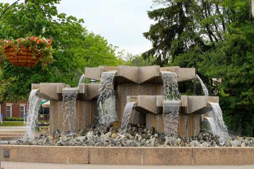 A modern stone fountain with cascading water, surrounded by greenery and flower baskets.