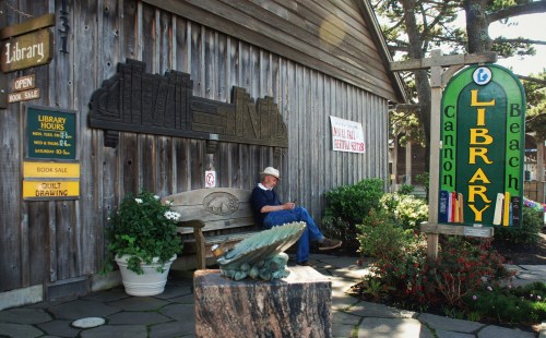 A man sits on a bench outside the Cannon Beach Library, surrounded by greenery and a colorful library sign.