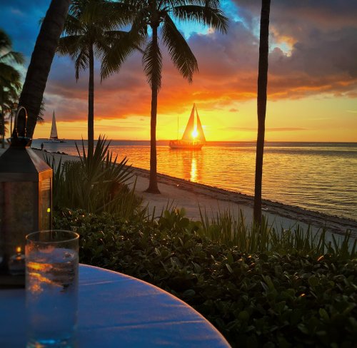 A serene sunset over the ocean with a sailboat in the distance, framed by palm trees and a table with a drink.