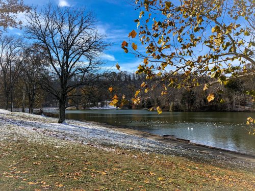 A serene lakeside scene with autumn leaves, a snowy shore, and a clear blue sky reflecting on the water.