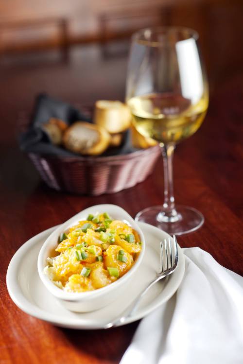 A creamy dish topped with green onions, served in a white bowl, alongside a glass of white wine and a basket of bread.