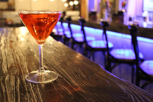 A vibrant cocktail in a martini glass sits on a wooden bar, with a softly lit background featuring blue bar stools.