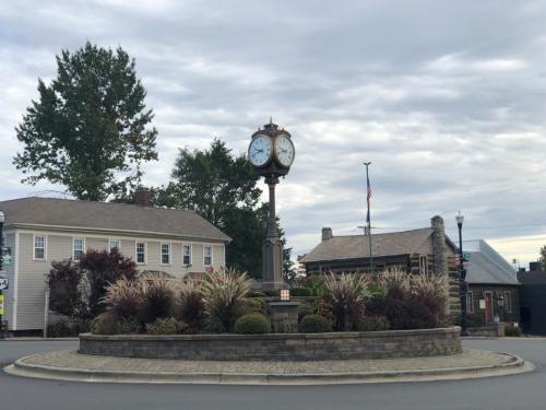 A clock tower stands in a roundabout surrounded by greenery and buildings under a cloudy sky.