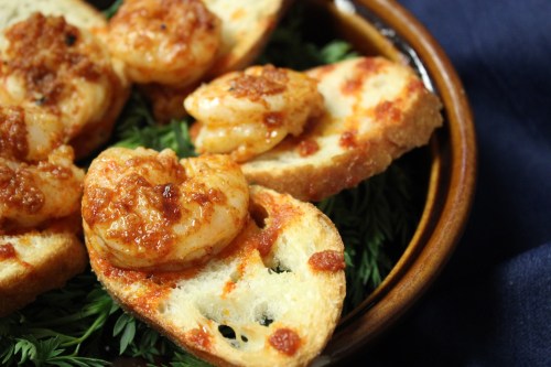 A close-up of toasted bread topped with shrimp in a savory sauce, served on a bed of greens in a brown dish.