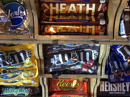 A display of various candy bars, including Heath, M&M's, Milky Way, Reese's, and Hershey's, in a wooden rack.