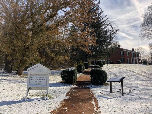 A snowy path leads to the Butler-Turner State Historic House, surrounded by trees and shrubs.